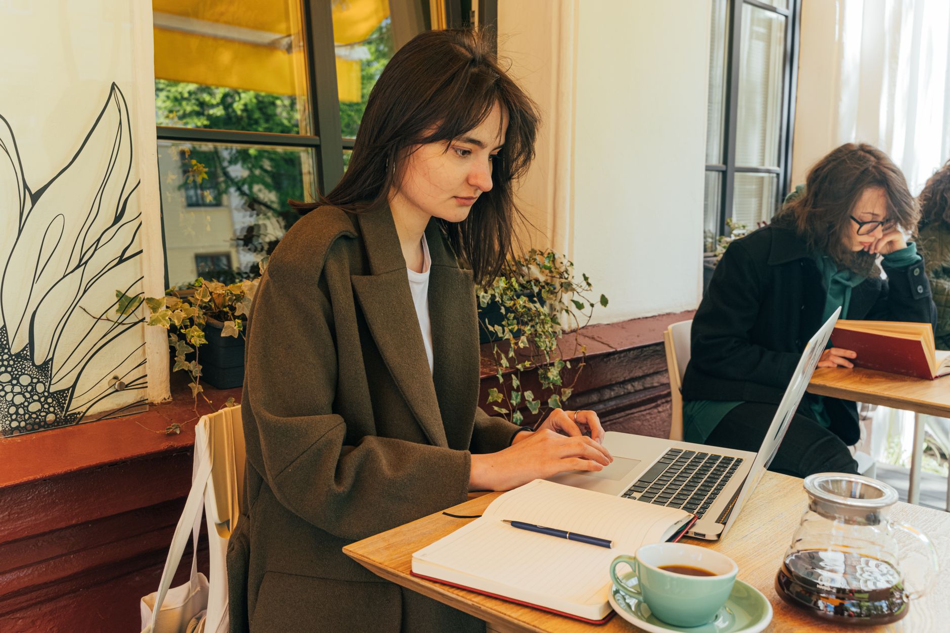 Une femme en manteau travaille sur un ordinateur portable à une table de café, avec un carnet ouvert, un stylo, une tasse et une carafe en verre.